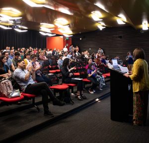 Auditorio con personas sentadas. Al frente una persona de pie en el podio hablando