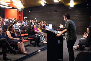 Auditorio con personas sentadas. Al frente una persona de pie en el podio hablando